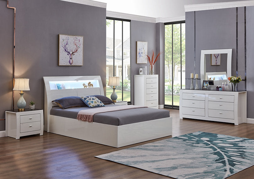 White bedroom furniture set with bed, nightstands, dresser, and mirror in a room with large windows.